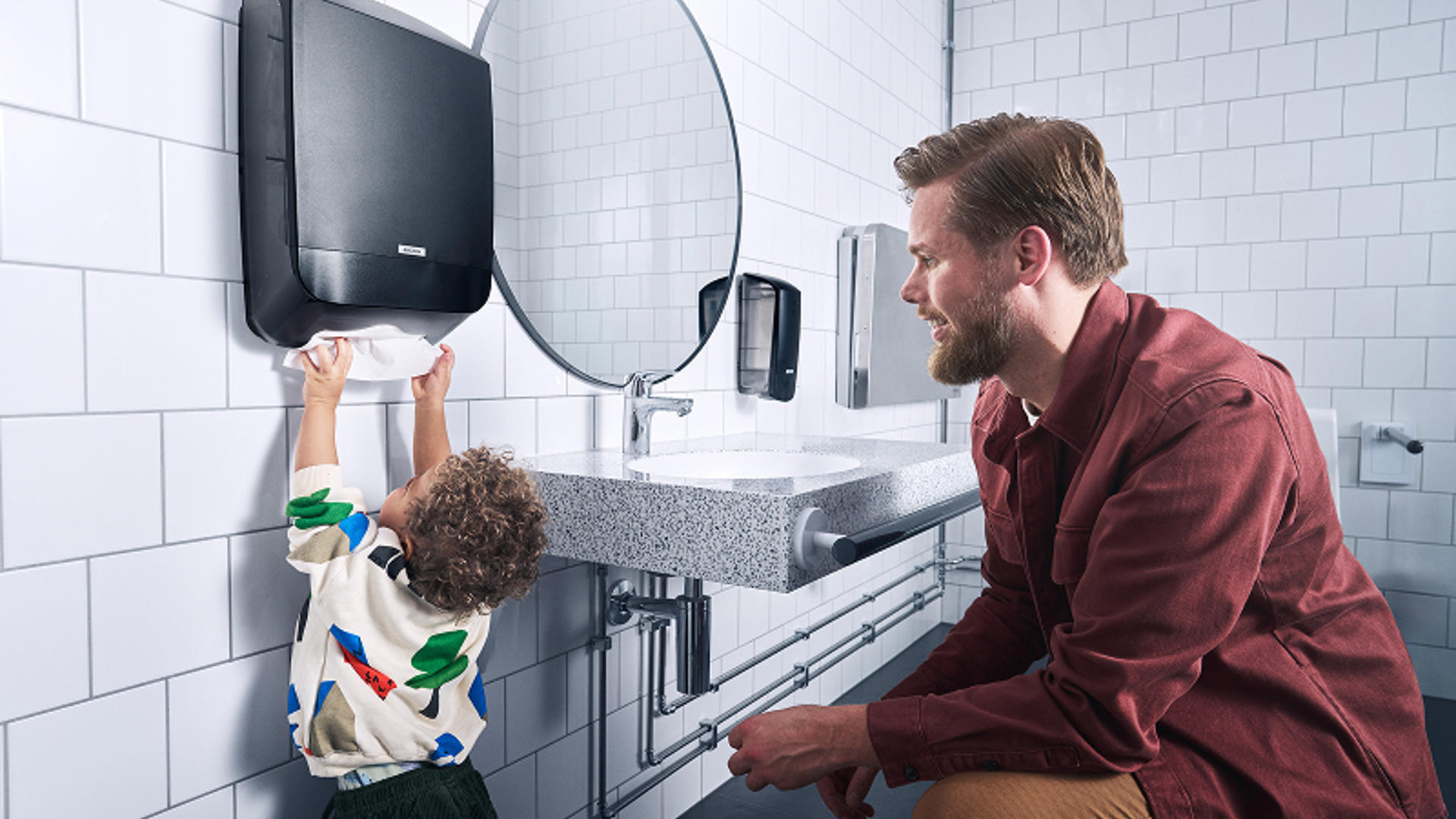Barn som når papper från en Katrin‑dispenser i ett offentligt tvättrum, som exempel på hur Katrin anpassar hygienlösningar efter användarmiljö.