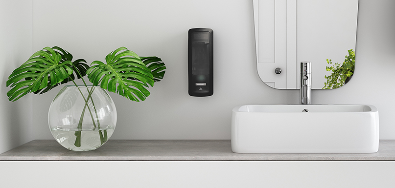 Et moderne badeværelse med en sort Katrin dispenser monteret på væggen ved siden af et hvidt håndvask med spejl. På bordpladen står en glasvase med grønne monstera-blade som dekoration.