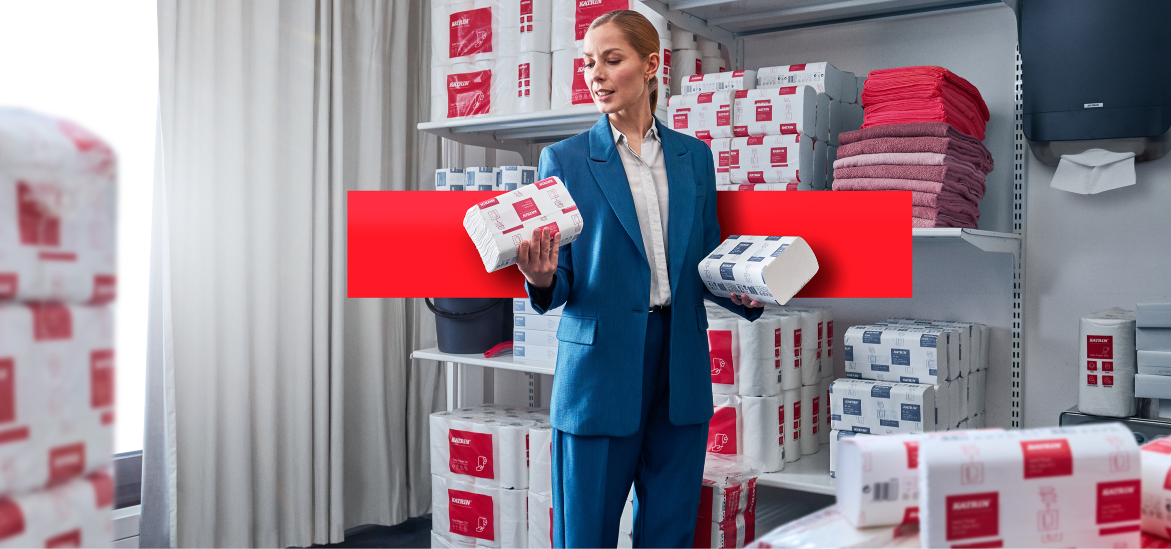 Kvinna håller förpackningar med Katrin mjukpapper i ett lager med staplade pappersprodukter från Katrin på hyllor.