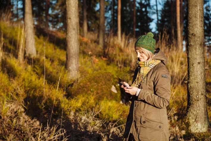 Nainen seisoo metsässä ja käyttää puhelimellaan Metsäverkko-mobiilia
