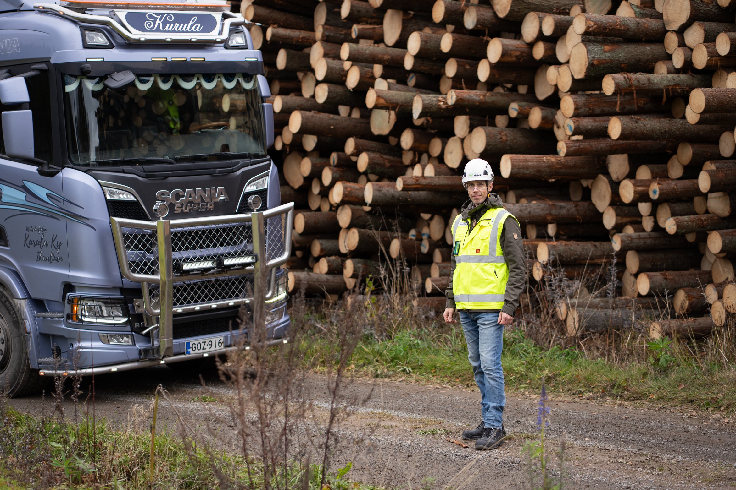 Ilkka Köntti står bredvid en timmerbil med en virkestrav i bakgrunden.