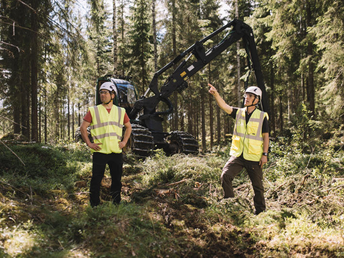 Skogsmaskinförare och skogsägare står framför maskinen och tittar på den omgivande skogen.