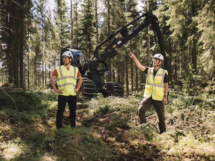 A forest machine operator and forest owner stand in front of the machine, looking at the surrounding forest.