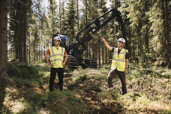 A forest machine operator and forest owner stand in front of the machine, looking at the surrounding forest.