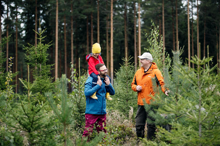 A child is sitting on the father’s shoulders, and accompanied by a forest specialist, they are looking at seedlings.