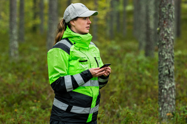 En skogsexpert står i skogen med telefon i hand och värderar den omgivande skogen.