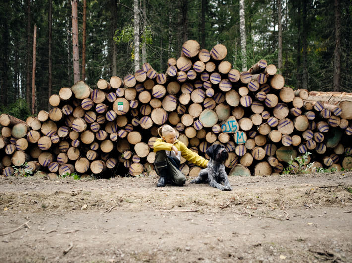A boy crouching down by a dog in front of a wood pile.