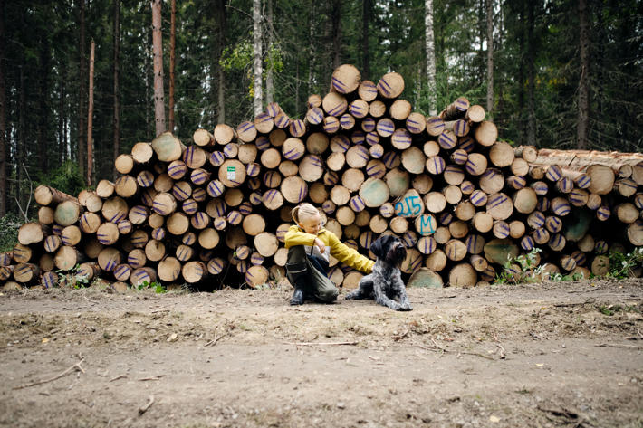 A boy crouching down by a dog in front of a wood pile.